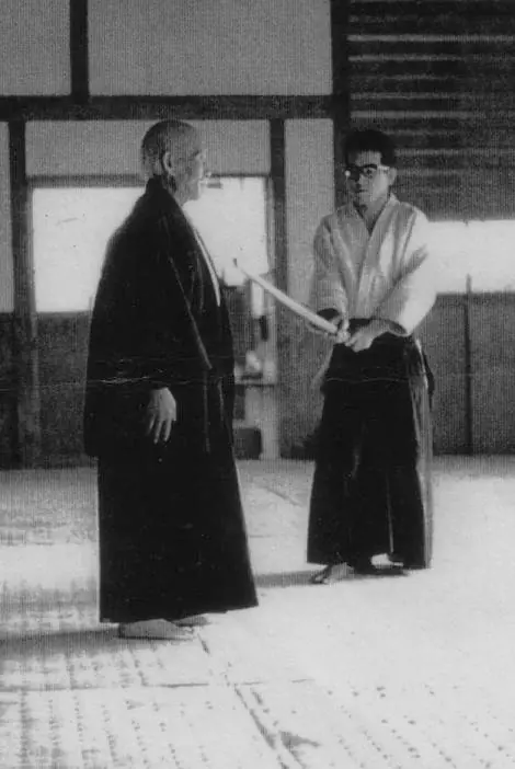Morihei Ueshiba âgé, en kimono sombre, face à son élève Yutaka Kurita qui tient un sabre de bois, dans un dojo en 1962